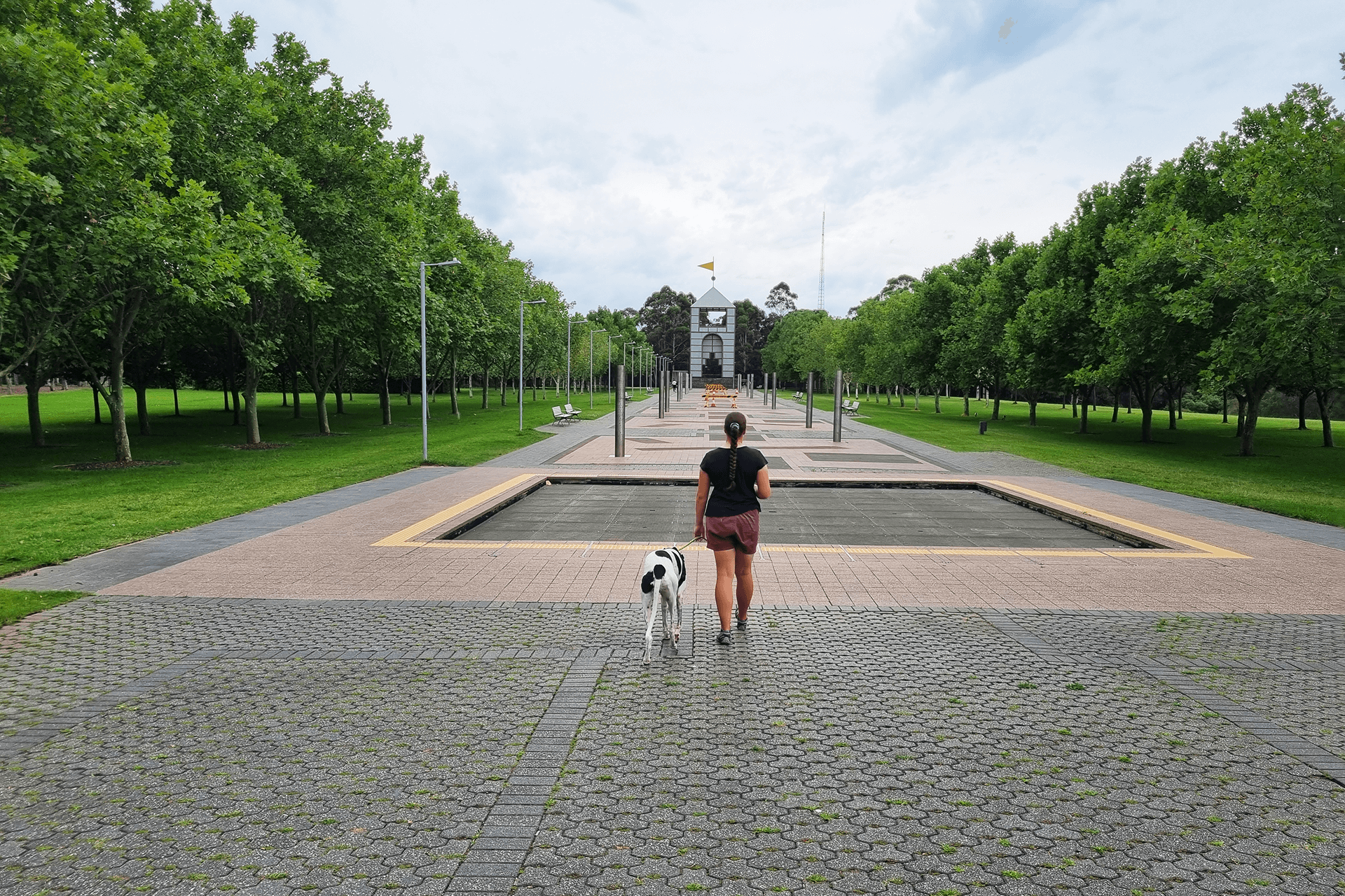 Person with dog facing paved walkway, lined with trees and grass on both sides, and the Treillage tower.