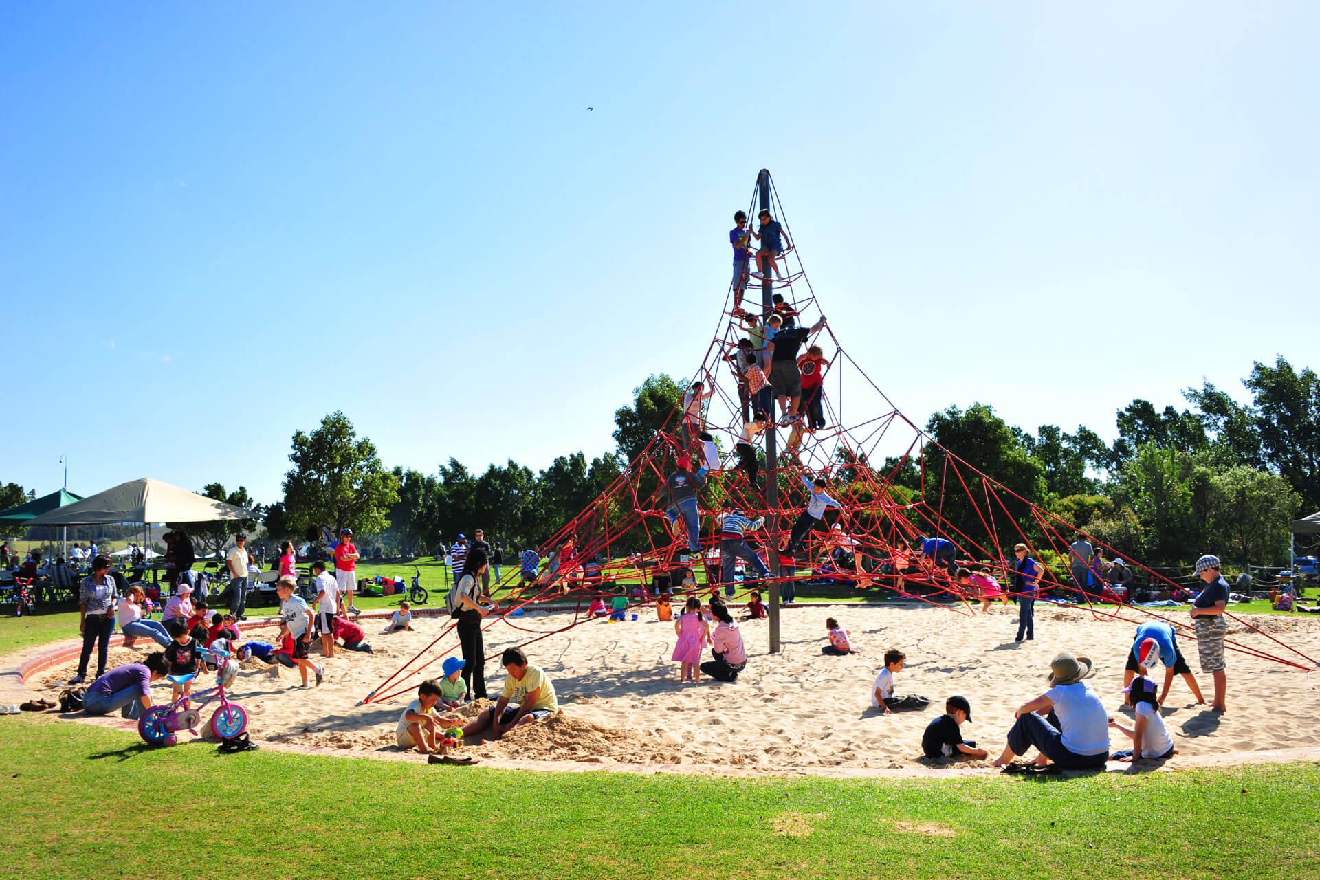 Many people playing on and around a rope climbing structure and sand pit.