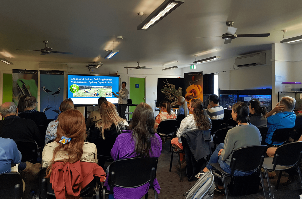 Multiple people seated in a room facing presenter and slideshow. Around the room are multiple posters of plants and animals, and glass aquaria.